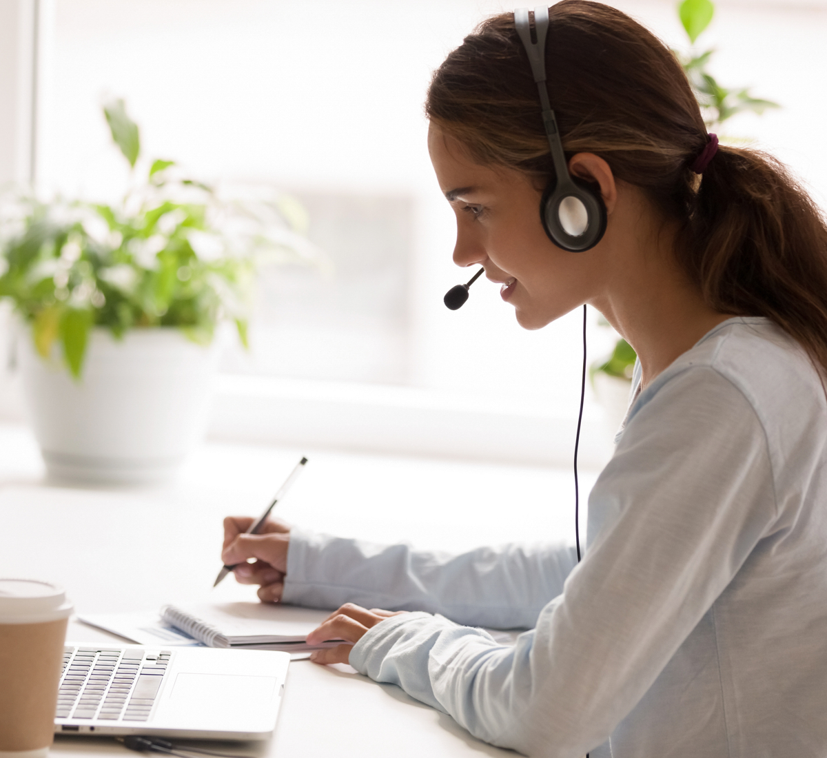 Side View Concentrated Millennial Mixed Race Woman Wearing Headset With Microphone, Looking At Laptop Screen, Listening Educational Webinar, Taking Notes, Getting Remote Online Education At Home.